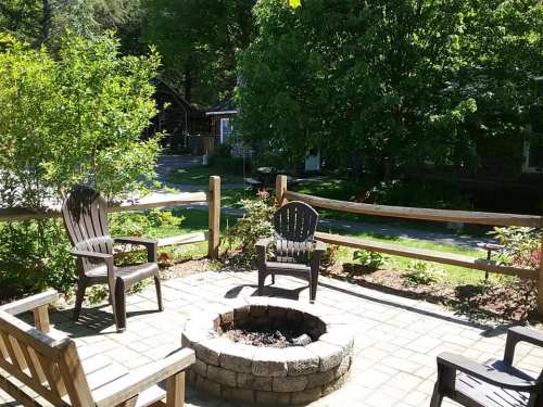 A cozy outdoor seating area with chairs around a stone fire pit, surrounded by greenery and a wooden fence.