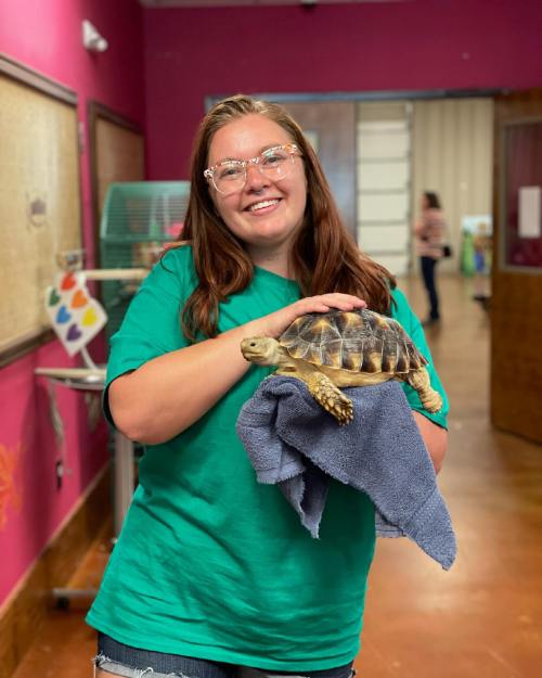 A smiling woman in a green shirt holds a tortoise wrapped in a towel in a colorful indoor space.