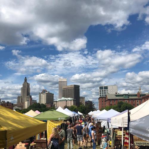 A bustling outdoor market with colorful tents, crowds of people, and a skyline of buildings under a blue sky with clouds.