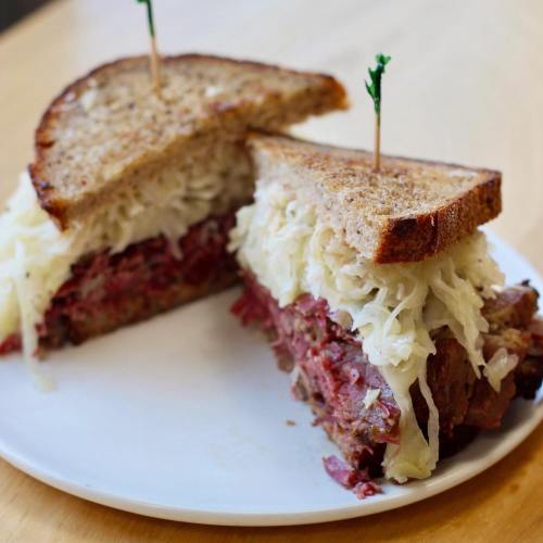 A close-up of a sandwich with layers of corned beef and sauerkraut, served on whole grain bread, garnished with a toothpick.
