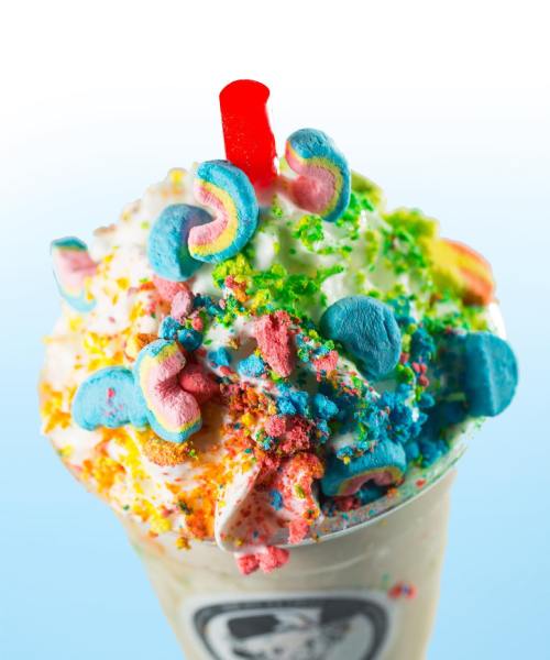 A colorful milkshake topped with whipped cream, rainbow sprinkles, and candy, featuring a red straw.
