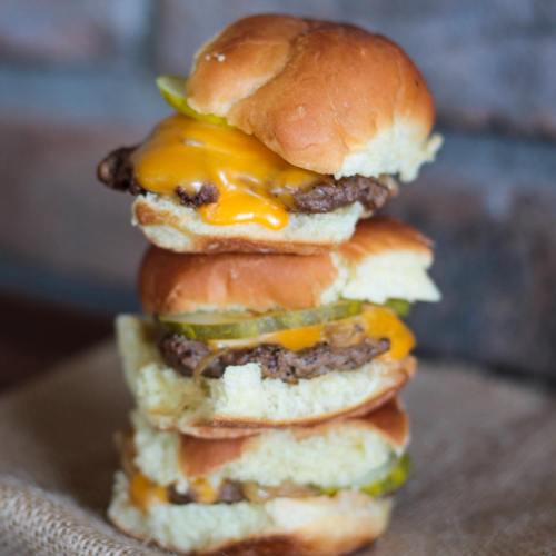 A stack of three mini burgers with melted cheese, pickles, and soft buns on a burlap surface.