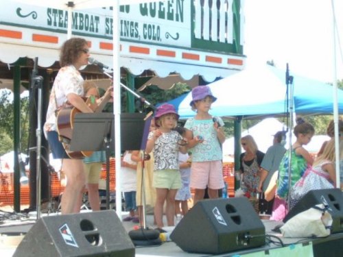 A performer with a guitar sings on stage with two children, all wearing colorful hats, at an outdoor festival.