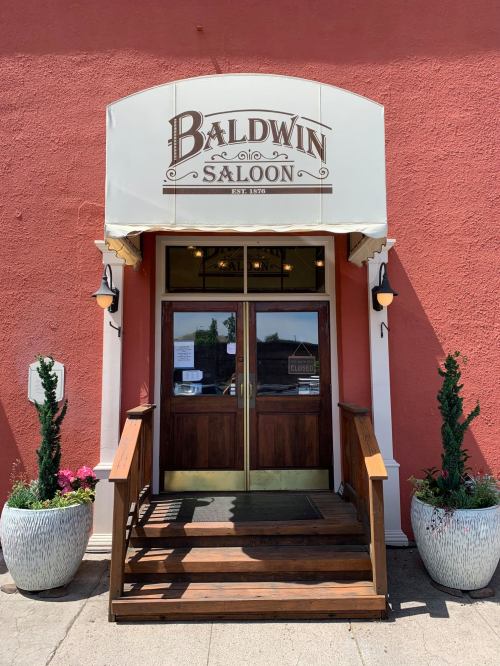 Entrance of Baldwin Saloon with a sign, wooden steps, and potted plants on either side against a red wall.