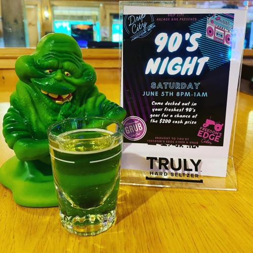 A green figurine next to a shot glass and a flyer for a '90s Night event at an arcade bar.