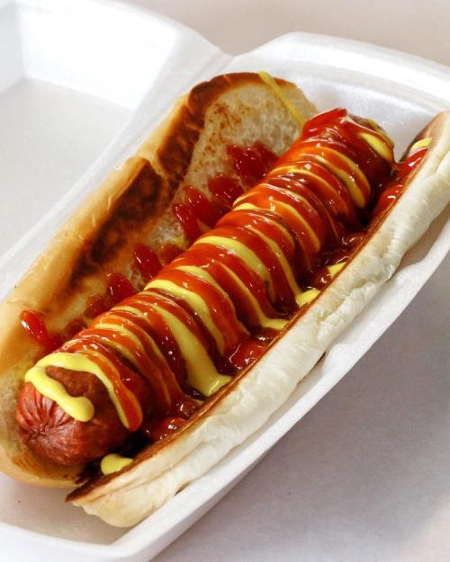 A hot dog in a bun topped with ketchup and mustard, served in a white takeout container.