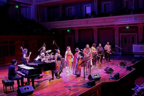 A diverse group of musicians performs on stage with instruments in a well-lit concert hall.
