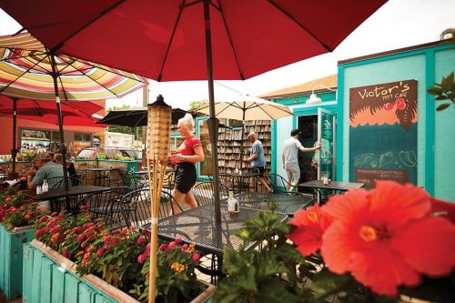 A vibrant outdoor café scene with colorful umbrellas, flowers, and people enjoying their time at tables.