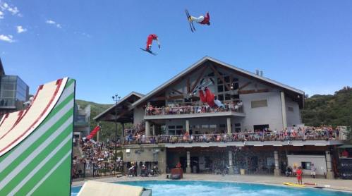 Skiers perform aerial tricks above a crowd at a water ramp event, with a building in the background under a clear blue sky.