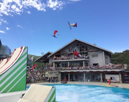 A skier performs a trick mid-air above a water ramp, with a crowd watching from a nearby building under a clear blue sky.