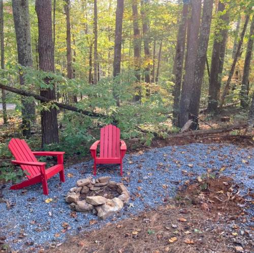 Two red Adirondack chairs beside a stone fire pit, surrounded by trees and autumn foliage.