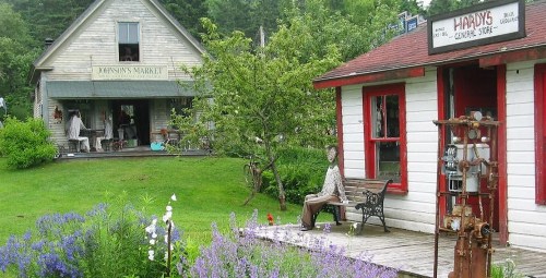 A quaint market and shop surrounded by greenery and flowers, featuring a bench and a decorative figure.