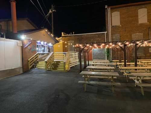 Outdoor seating area at night with picnic tables and string lights, next to a food stand in a brick building setting.