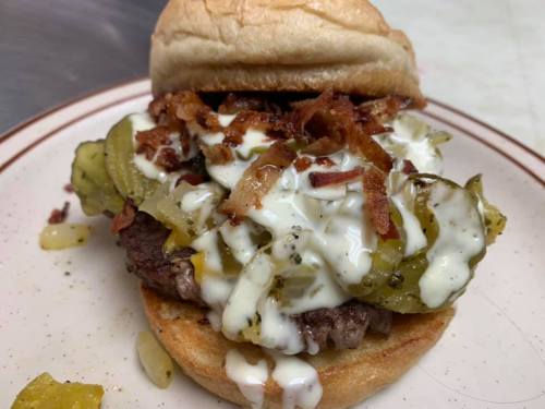 A close-up of a burger topped with pickles, bacon, and creamy sauce, served on a toasted bun.