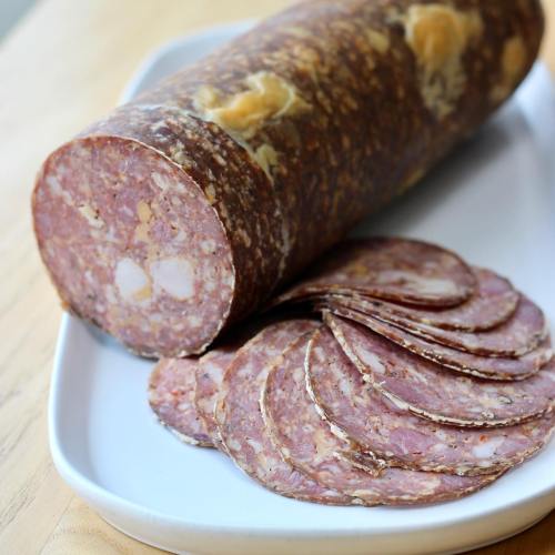 A sliced log of salami on a white plate, showcasing thin, round slices next to the whole piece.