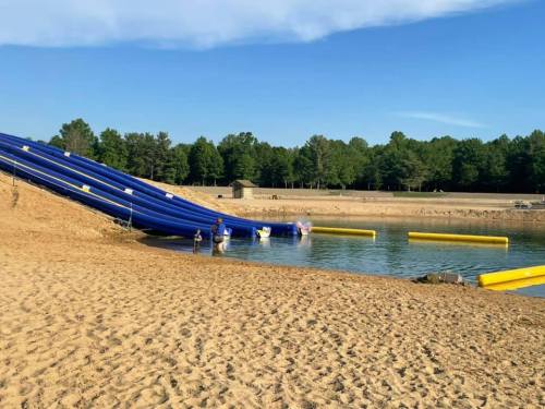 A sandy beach area with a large blue water slide leading into a calm lake, surrounded by trees and clear skies.