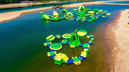 A colorful inflatable water park with various structures on a lake, surrounded by sandy shores and greenery.