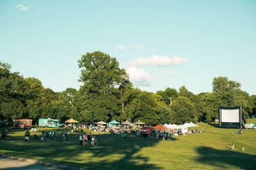 A park scene with people gathered around food tents and a large outdoor movie screen under a clear blue sky.