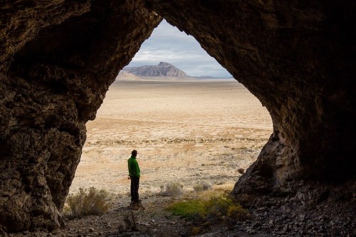 A person stands in a cave, gazing out at a vast desert landscape and distant mountains under a cloudy sky.