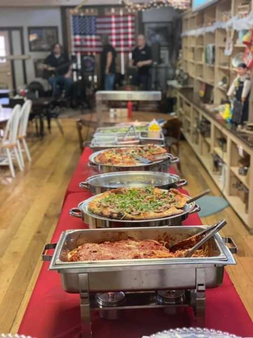 A buffet table with various dishes, including pasta and pizza, set in a cozy room with an American flag in the background.