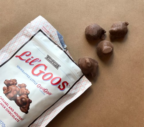 A bag of Lil' Goos candy with a few pieces scattered on a brown surface. The candy is chocolate-covered caramel and nougat.