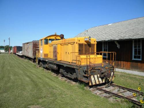 A yellow diesel locomotive parked beside a wooden building, with freight cars on the tracks under a clear blue sky.