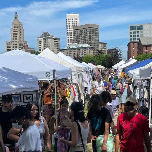 A bustling outdoor market with tents, vendors, and a crowd, set against a city skyline on a sunny day.