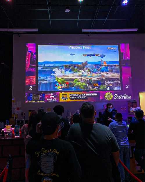 A crowd watches a large screen displaying a gaming tournament match, with scores and logos visible.