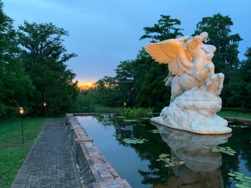 A white statue of a winged figure stands by a tranquil pond, surrounded by lush greenery and a sunset in the background.