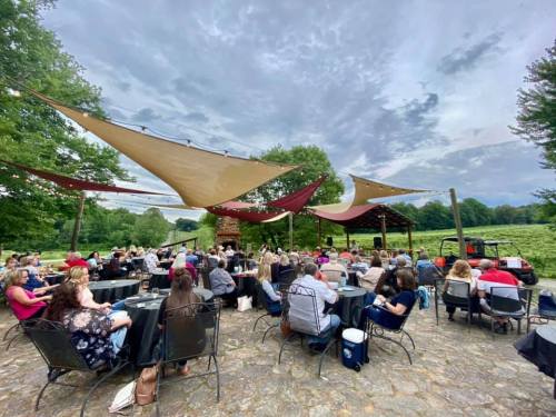 A large outdoor gathering under shaded canopies, with people seated at tables, surrounded by greenery and a cloudy sky.