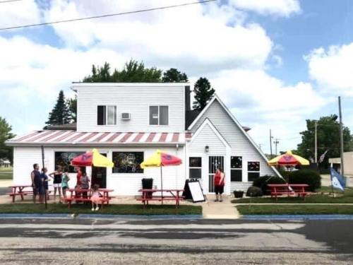 A small white restaurant with a red roof, outdoor seating, and colorful umbrellas, surrounded by trees and a blue sky.
