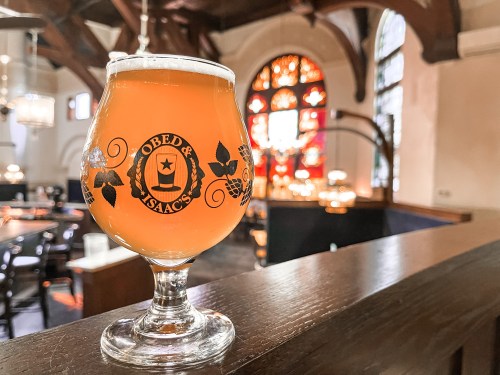 A glass of amber beer sits on a wooden ledge, with a cozy restaurant interior and stained glass windows in the background.