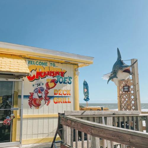 A colorful sign for Crabby Joe's Deck Grill, featuring a crab and a fish, with a beach view and blue sky.