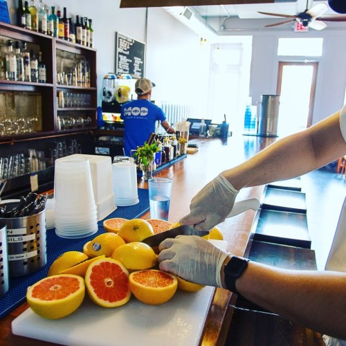 A person in gloves slices citrus fruits on a bar counter, with a bartender preparing drinks in the background.
