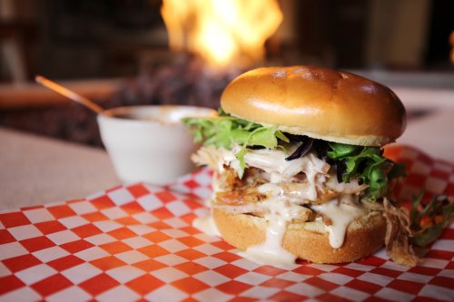 A delicious burger with shredded chicken, lettuce, and sauce, served on a checkered paper with a side of dipping sauce.