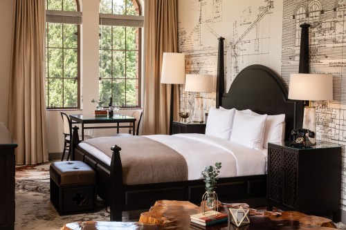 A stylish hotel room featuring a bed, desk, and large windows with natural light, accented by elegant decor.