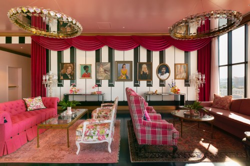 A stylish lounge with pink sofas, floral decor, and framed portraits against a striped wall, featuring elegant chandeliers.