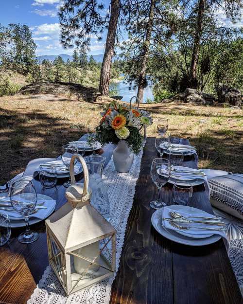 A beautifully set outdoor dining table with flowers, glassware, and a lantern, overlooking a scenic river and trees.