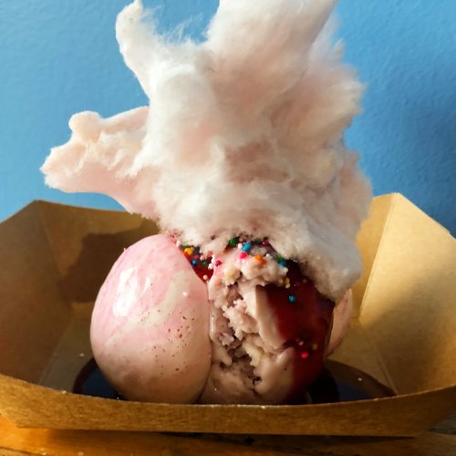 A dessert featuring pink ice cream topped with cotton candy, sprinkles, and a drizzle of chocolate sauce in a paper bowl.