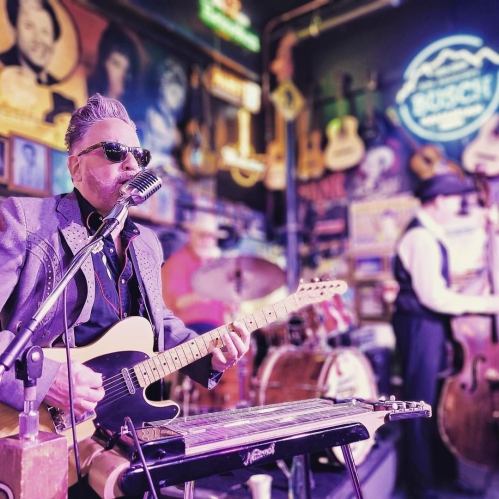 A musician in sunglasses plays guitar and sings into a vintage microphone, with a drummer and another musician in the background.