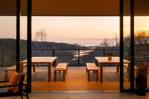 Modern outdoor seating area with wooden tables, overlooking a serene sunset view by the water.