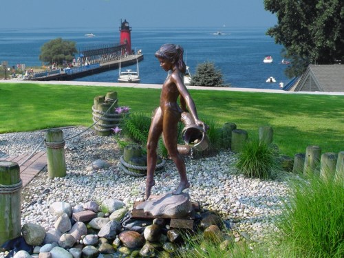 A bronze statue of a girl holding a water jug stands on a stone base, overlooking a lake and lighthouse in the background.