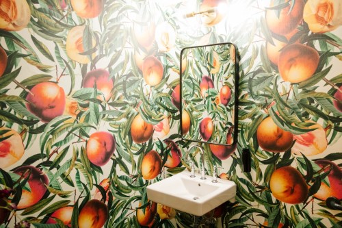 A bathroom with a sink and a mirror, surrounded by vibrant fruit-patterned wallpaper featuring peaches and leaves.