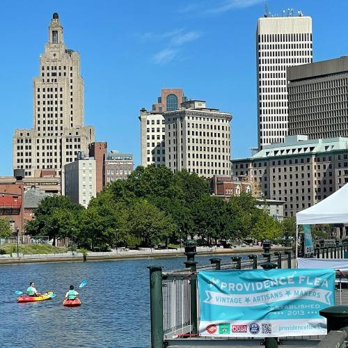 Two kayakers paddle on a river with city buildings in the background, featuring a banner for the Providence Flea.