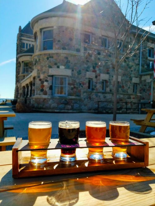 A wooden flight of four craft beers sits on a table, with a stone building and sunlight in the background.