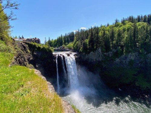 A stunning waterfall cascades down rocky cliffs, surrounded by lush green trees under a clear blue sky.