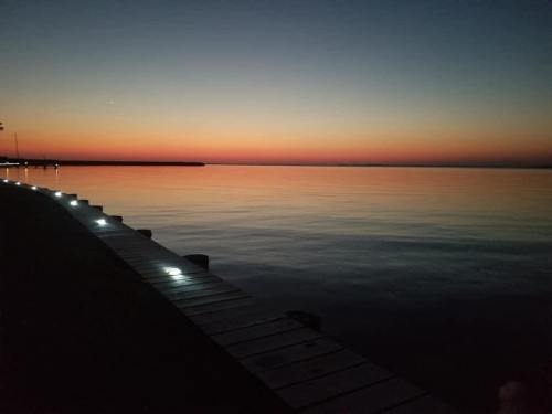 A serene sunset over calm waters, with a wooden pier illuminated by soft lights along the edge.