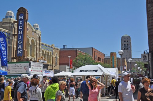 A bustling street fair with people, tents, and buildings, including a theater, under a clear blue sky.