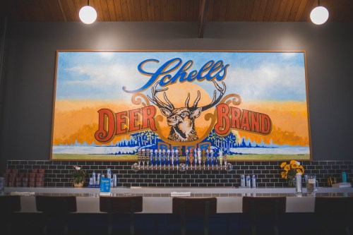 A colorful mural of a deer and the text "Schell's Deer Brand Beer" on a wall above a bar with taps and decor.