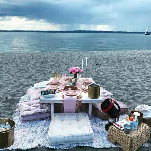 A beach picnic setup with a table, pink decor, cushions, and candles, overlooking a calm sea under cloudy skies.